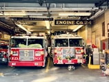 Two red fire trucks are parked inside a fire station garage. The station has signage indicating 'Engine Co. 24' and 'Ladder Co. 5'. The trucks are adorned with American flags and various firefighting equipment. The setting includes visible pipes and lighting fixtures on the ceiling.