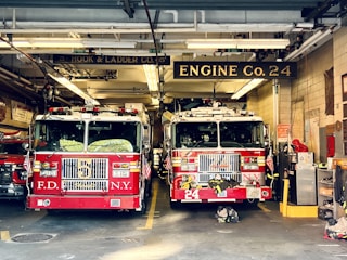 Two red fire trucks are parked inside a fire station garage. The station has signage indicating 'Engine Co. 24' and 'Ladder Co. 5'. The trucks are adorned with American flags and various firefighting equipment. The setting includes visible pipes and lighting fixtures on the ceiling.