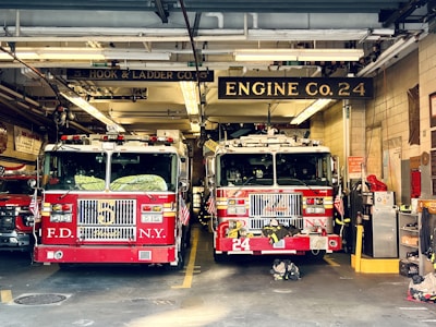 Two red fire trucks are parked inside a fire station garage. The station has signage indicating 'Engine Co. 24' and 'Ladder Co. 5'. The trucks are adorned with American flags and various firefighting equipment. The setting includes visible pipes and lighting fixtures on the ceiling.