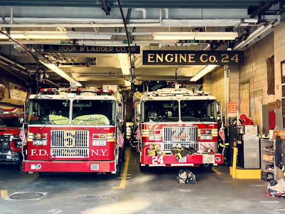 Two red fire trucks are parked inside a fire station garage. The station has signage indicating 'Engine Co. 24' and 'Ladder Co. 5'. The trucks are adorned with American flags and various firefighting equipment. The setting includes visible pipes and lighting fixtures on the ceiling.