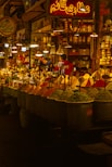 A vibrant spice market with a variety of colorful spices piled in mounds. The warm, golden lighting gives a cozy atmosphere. Various signs in a different language are visible, and a few people are engaged in conversation or transactions.