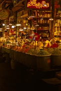 A vibrant spice market with a variety of colorful spices piled in mounds. The warm, golden lighting gives a cozy atmosphere. Various signs in a different language are visible, and a few people are engaged in conversation or transactions.