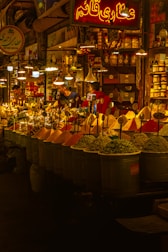 A vibrant spice market with a variety of colorful spices piled in mounds. The warm, golden lighting gives a cozy atmosphere. Various signs in a different language are visible, and a few people are engaged in conversation or transactions.