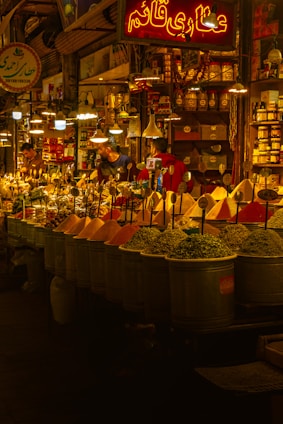 A vibrant spice market with a variety of colorful spices piled in mounds. The warm, golden lighting gives a cozy atmosphere. Various signs in a different language are visible, and a few people are engaged in conversation or transactions.