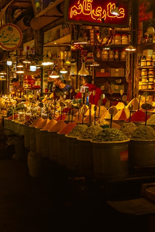 A vibrant spice market with a variety of colorful spices piled in mounds. The warm, golden lighting gives a cozy atmosphere. Various signs in a different language are visible, and a few people are engaged in conversation or transactions.