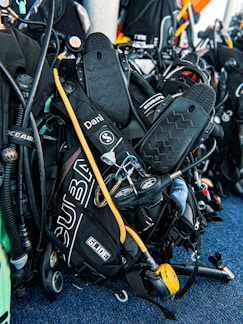 A collection of scuba diving equipment, including flippers, breathing regulators, air cylinders, and gauges, is arranged on a textured blue surface. The gear has various labels and branding, including 'Scubapro' and 'Oceanic.'