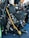 A collection of scuba diving equipment, including flippers, breathing regulators, air cylinders, and gauges, is arranged on a textured blue surface. The gear has various labels and branding, including 'Scubapro' and 'Oceanic.'