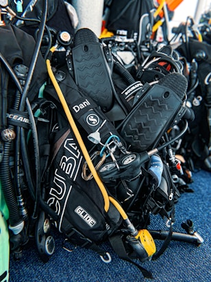 A collection of scuba diving equipment, including flippers, breathing regulators, air cylinders, and gauges, is arranged on a textured blue surface. The gear has various labels and branding, including 'Scubapro' and 'Oceanic.'