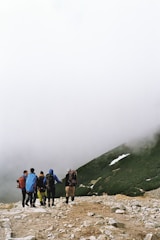 A group of five hikers are walking together on a rocky trail covered with stones, heading towards a foggy and mountainous area. They are dressed in outdoor gear and carrying backpacks. The surrounding landscape includes patches of greenery and a steep slope partially covered in snow. The background is dominated by dense fog or mist, creating a mysterious and adventurous atmosphere.