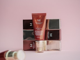 A red tube of BB cream is positioned in front of a series of stacked boxes with a reflective finish. The tube has the brand name and product details printed on it. The background is a soft pink color, adding a warm and gentle aesthetic to the scene.