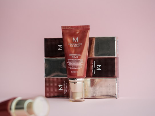 A red tube of BB cream is positioned in front of a series of stacked boxes with a reflective finish. The tube has the brand name and product details printed on it. The background is a soft pink color, adding a warm and gentle aesthetic to the scene.