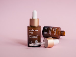 Three brown dropper bottles are displayed on a soft pink background. One bottle stands upright with a closed dropper, while another lies on its side, with the dropper removed and resting in front. The bottles have labels suggesting they contain a serum product.