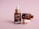 Three brown dropper bottles are displayed on a soft pink background. One bottle stands upright with a closed dropper, while another lies on its side, with the dropper removed and resting in front. The bottles have labels suggesting they contain a serum product.