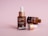 Three brown dropper bottles are displayed on a soft pink background. One bottle stands upright with a closed dropper, while another lies on its side, with the dropper removed and resting in front. The bottles have labels suggesting they contain a serum product.