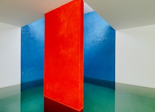A minimalist interior space features a striking vertical red wall dividing two contrasting sections. One side exhibits a deep blue wall, while the other shows a light white wall. The flooring reflects the walls' colors, blending green and blue hues under soft lighting.