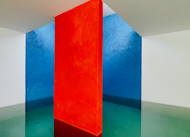 A minimalist interior space features a striking vertical red wall dividing two contrasting sections. One side exhibits a deep blue wall, while the other shows a light white wall. The flooring reflects the walls' colors, blending green and blue hues under soft lighting.