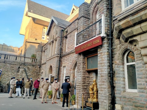 Several uniformed police officers and a few people stand outside a historic stone building with arched windows and a sign that reads 'Police Assistance Room'. The structure features classic architectural details and a statue placed near the entrance. The scene is set in daylight, with a clear sky visible in the background.