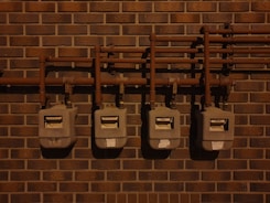 Four utility meters mounted on a brick wall, connected by a series of pipes and fittings. The scene is dimly lit, casting shadows on the wall and the meters.