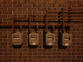 Four utility meters mounted on a brick wall, connected by a series of pipes and fittings. The scene is dimly lit, casting shadows on the wall and the meters.