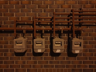 Four utility meters mounted on a brick wall, connected by a series of pipes and fittings. The scene is dimly lit, casting shadows on the wall and the meters.