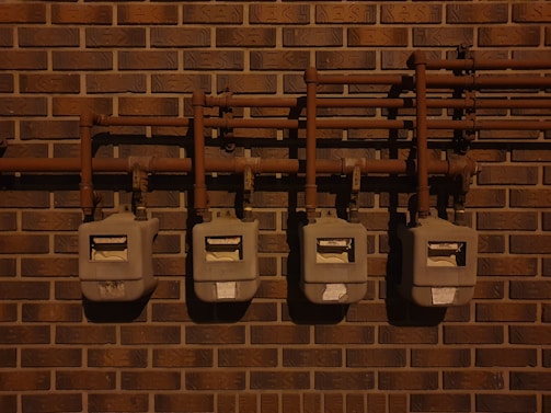 Four utility meters mounted on a brick wall, connected by a series of pipes and fittings. The scene is dimly lit, casting shadows on the wall and the meters.