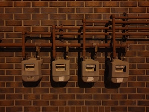 Four utility meters mounted on a brick wall, connected by a series of pipes and fittings. The scene is dimly lit, casting shadows on the wall and the meters.