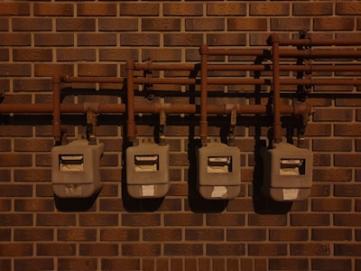 Four utility meters mounted on a brick wall, connected by a series of pipes and fittings. The scene is dimly lit, casting shadows on the wall and the meters.