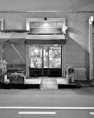 A black and white image of a storefront with an awning displaying the name 'ERBA BRUSCA'. The entrance includes two double doors with glass panels. The interior is faintly visible, showing some shelves and lighting inside. There are potted plants and a wooden bench outside, along with a small ramp leading to the door. The street in front of the store is empty.