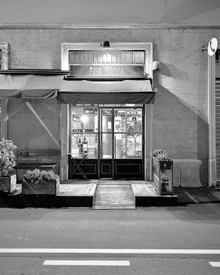 A black and white image of a storefront with an awning displaying the name 'ERBA BRUSCA'. The entrance includes two double doors with glass panels. The interior is faintly visible, showing some shelves and lighting inside. There are potted plants and a wooden bench outside, along with a small ramp leading to the door. The street in front of the store is empty.