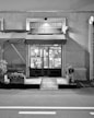 A black and white image of a storefront with an awning displaying the name 'ERBA BRUSCA'. The entrance includes two double doors with glass panels. The interior is faintly visible, showing some shelves and lighting inside. There are potted plants and a wooden bench outside, along with a small ramp leading to the door. The street in front of the store is empty.