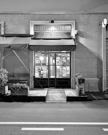 A black and white image of a storefront with an awning displaying the name 'ERBA BRUSCA'. The entrance includes two double doors with glass panels. The interior is faintly visible, showing some shelves and lighting inside. There are potted plants and a wooden bench outside, along with a small ramp leading to the door. The street in front of the store is empty.