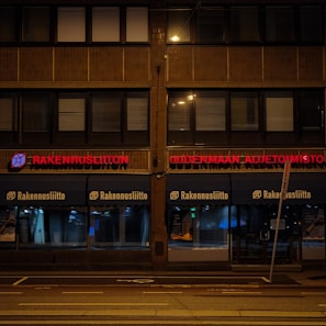 A building facade at night with illuminated red signage displaying 'RAKENNUSLIITON UUDENMAAN ALUETOIMISTO'. The building has a row of large windows with blinds, and the sidewalk in front is empty and dimly lit by street lights.