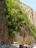 A narrow road bordered by steep, rocky cliffs and lush green vegetation. Several vehicles, including cars and a white bus, are parked along the roadside. The scene conveys a sense of adventure and nature's majesty as the road winds through the cliffside.