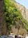 A narrow road bordered by steep, rocky cliffs and lush green vegetation. Several vehicles, including cars and a white bus, are parked along the roadside. The scene conveys a sense of adventure and nature's majesty as the road winds through the cliffside.