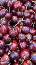 A close-up view of a pile of fresh, ripe cherries with glossy, deep red skins and green stems. Each cherry is plump and clustered close together, creating a lush and vibrant texture.