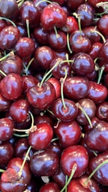 A close-up view of a pile of fresh, ripe cherries with glossy, deep red skins and green stems. Each cherry is plump and clustered close together, creating a lush and vibrant texture.