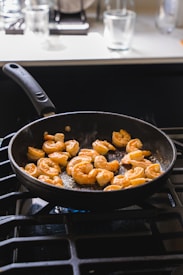A black frying pan is placed on a stovetop, containing several pieces of shrimp being cooked. The surrounding kitchen area is slightly blurred, with glass cups visible in the background.