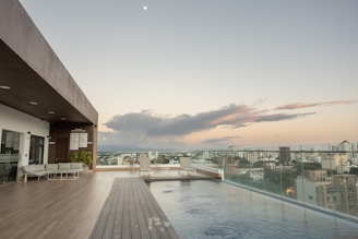 A rooftop infinity pool overlooks a cityscape at sunset, with the sky displaying hues of orange and purple. The pool area features a modern design with wooden decking, lounge chairs, and a canopy-covered seating area to the left. Glass railings provide an unobstructed view of the city buildings and the distant mountains.