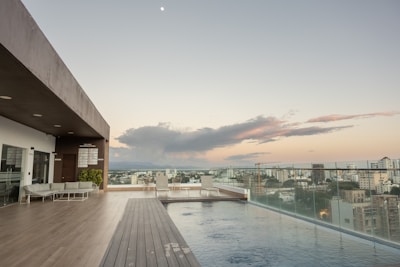 A rooftop infinity pool overlooks a cityscape at sunset, with the sky displaying hues of orange and purple. The pool area features a modern design with wooden decking, lounge chairs, and a canopy-covered seating area to the left. Glass railings provide an unobstructed view of the city buildings and the distant mountains.