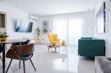A modern living room with a minimalistic design featuring a large window covered by sheer white curtains. The room includes a stylish yellow armchair, a green sofa, and a sleek wooden dining table with matching chairs. There is a wall-mounted TV, decorative plants, and wall art, enhancing the aesthetic. The floors are shiny and white, contributing to the spacious and bright atmosphere.