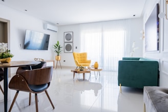 A modern living room with a minimalistic design featuring a large window covered by sheer white curtains. The room includes a stylish yellow armchair, a green sofa, and a sleek wooden dining table with matching chairs. There is a wall-mounted TV, decorative plants, and wall art, enhancing the aesthetic. The floors are shiny and white, contributing to the spacious and bright atmosphere.