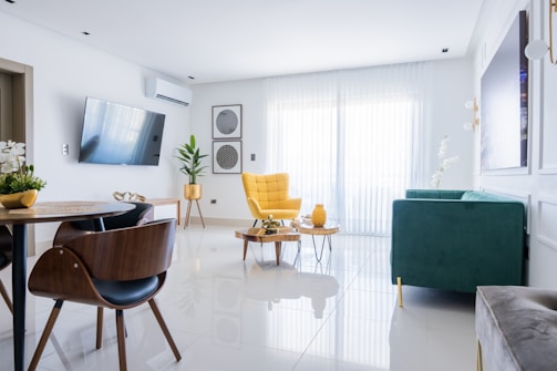 A modern living room with a minimalistic design featuring a large window covered by sheer white curtains. The room includes a stylish yellow armchair, a green sofa, and a sleek wooden dining table with matching chairs. There is a wall-mounted TV, decorative plants, and wall art, enhancing the aesthetic. The floors are shiny and white, contributing to the spacious and bright atmosphere.
