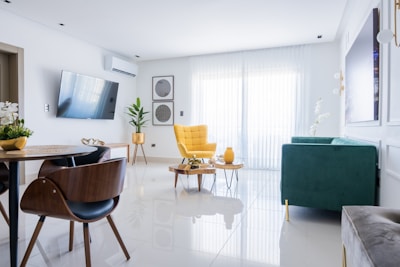 A modern living room with a minimalistic design featuring a large window covered by sheer white curtains. The room includes a stylish yellow armchair, a green sofa, and a sleek wooden dining table with matching chairs. There is a wall-mounted TV, decorative plants, and wall art, enhancing the aesthetic. The floors are shiny and white, contributing to the spacious and bright atmosphere.