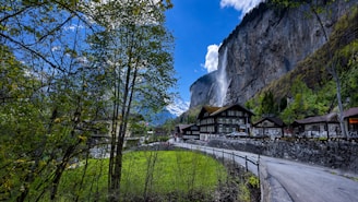 A picturesque village nestled in a valley with a dramatic waterfall cascading down a steep cliff. Quaint wooden houses are surrounded by lush greenery, and a narrow road winds through the landscape. Snow-capped mountains rise in the background under a bright blue sky adorned with fluffy clouds.
