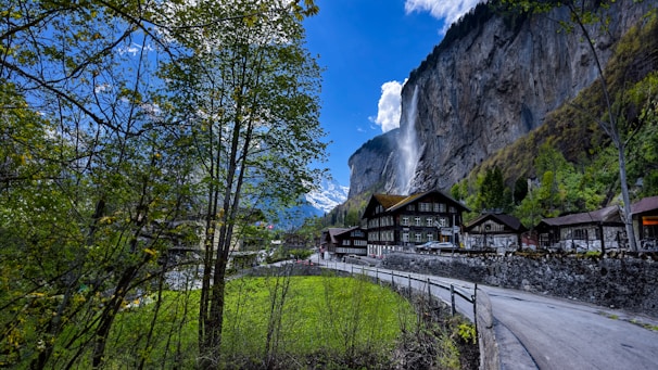 A picturesque village nestled in a valley with a dramatic waterfall cascading down a steep cliff. Quaint wooden houses are surrounded by lush greenery, and a narrow road winds through the landscape. Snow-capped mountains rise in the background under a bright blue sky adorned with fluffy clouds.