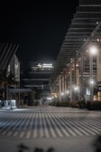 An urban night scene featuring a modern architectural facade with geometric designs and illuminated by soft lighting. The area appears quiet and empty, with a wide, striped pathway leading into the scene. A large building with 'DP World' signage is prominent in the background.