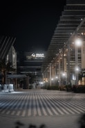 An urban night scene featuring a modern architectural facade with geometric designs and illuminated by soft lighting. The area appears quiet and empty, with a wide, striped pathway leading into the scene. A large building with 'DP World' signage is prominent in the background.