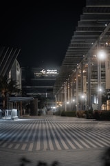 An urban night scene featuring a modern architectural facade with geometric designs and illuminated by soft lighting. The area appears quiet and empty, with a wide, striped pathway leading into the scene. A large building with 'DP World' signage is prominent in the background.