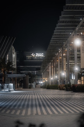 An urban night scene featuring a modern architectural facade with geometric designs and illuminated by soft lighting. The area appears quiet and empty, with a wide, striped pathway leading into the scene. A large building with 'DP World' signage is prominent in the background.