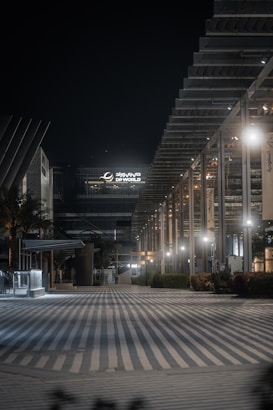 An urban night scene featuring a modern architectural facade with geometric designs and illuminated by soft lighting. The area appears quiet and empty, with a wide, striped pathway leading into the scene. A large building with 'DP World' signage is prominent in the background.